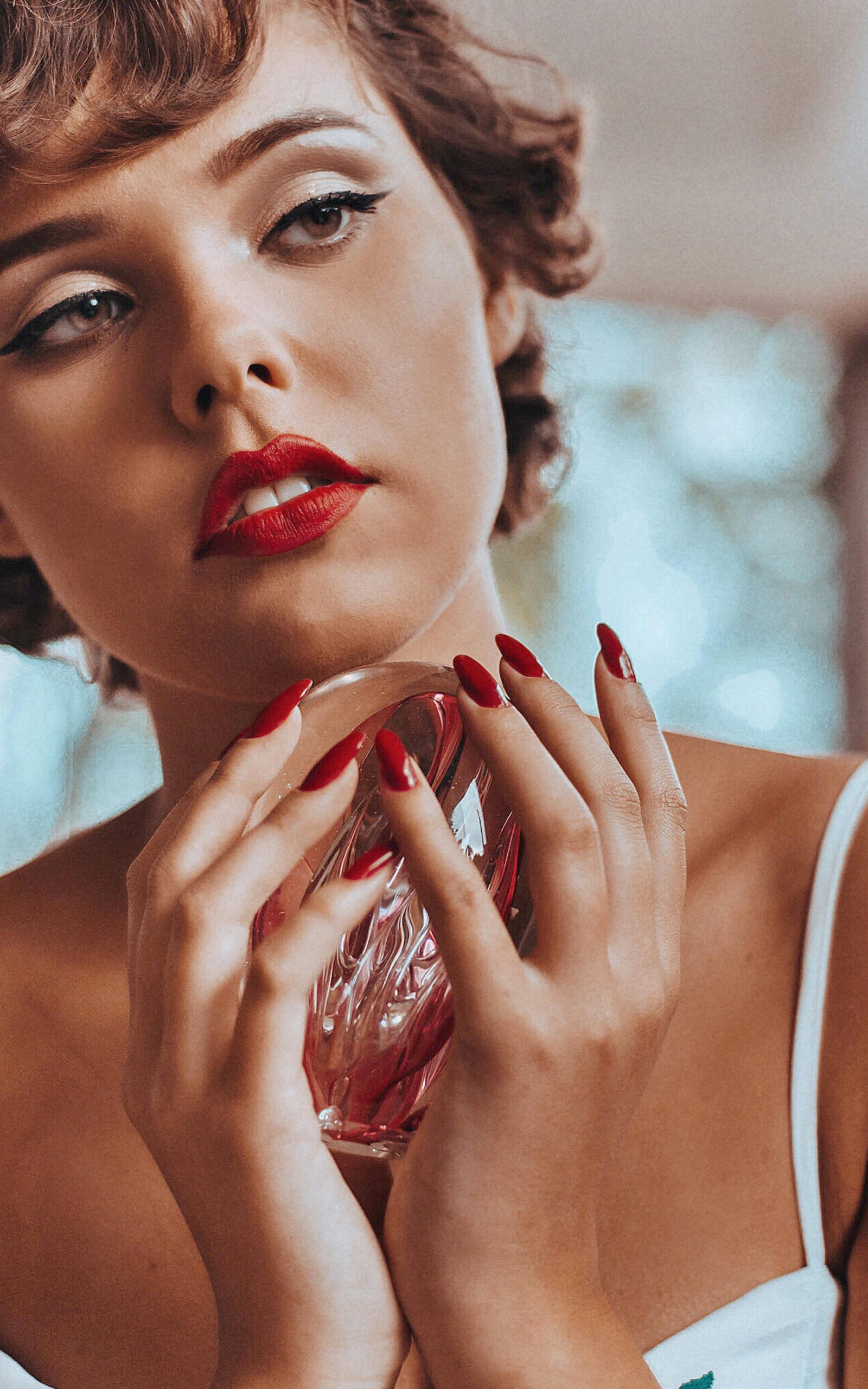 Elegant woman with red lipstick and nail polish holding a perfume bottle, portraying glamour.